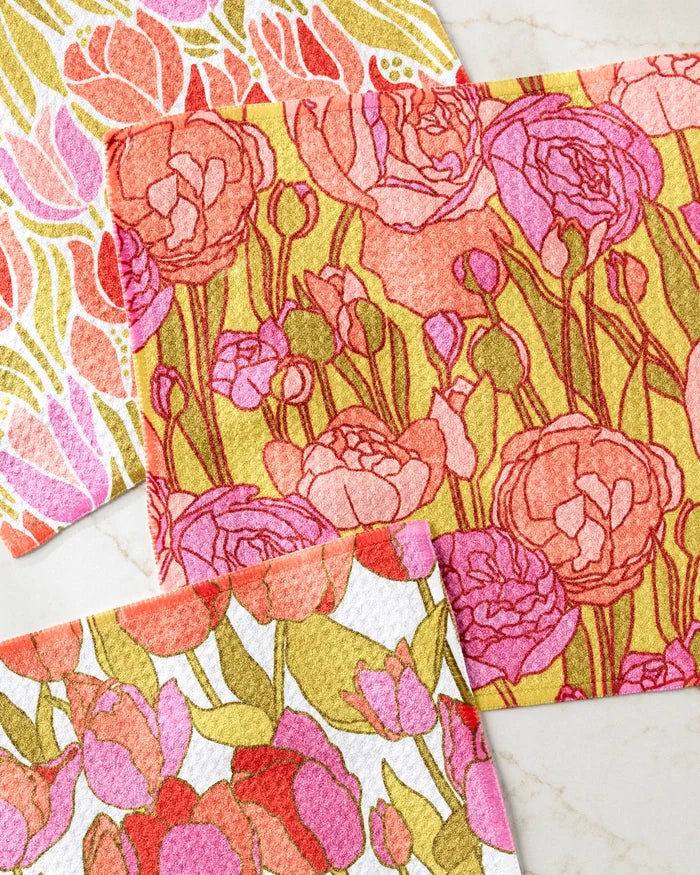 The Sidney dishcloth set by Amy MacCready features three rectangular cloths with vibrant floral and leaf patterns in pink, red, orange, and yellow. Made from recycled materials, they’re displayed on a light marble surface.