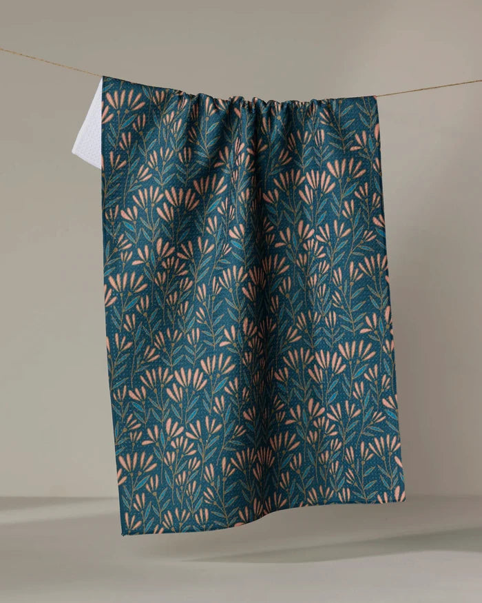 The Sirocco recycled kitchen towel by Amy MacCready, in dark blue with light pink and green leafy floral patterns, hangs over a clothesline against a simple, light background.