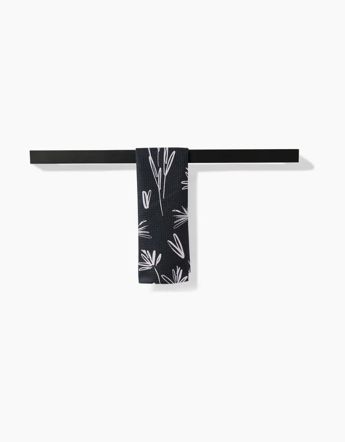 A Sketchbook Dust towel by Toboggan Avenue, crafted from soft Geoweave fabric, is neatly draped over a dark metal bar mounted on a white wall. The navy blue towel features a white floral pattern in the center.