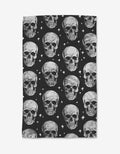 The GEOMETRY Skulls tea towel is rectangular, made from recycled materials, and features a repeating pattern of realistic gray skulls on black with small white star-like accents.