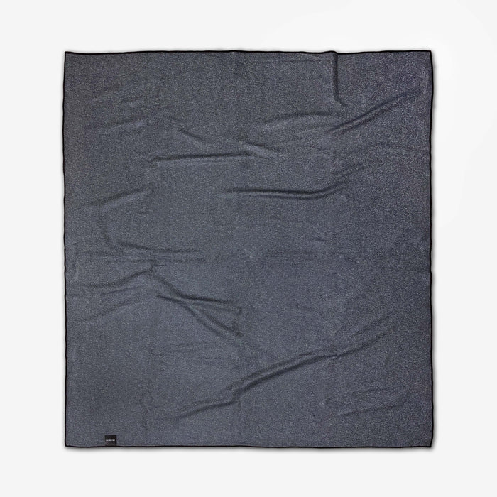 The GEOMETRY Sky Party beach blanket is large, dark gray, rectangular, and quick-drying with visible creases. It is laid flat on a white background and features a small black tag attached to one bottom corner.