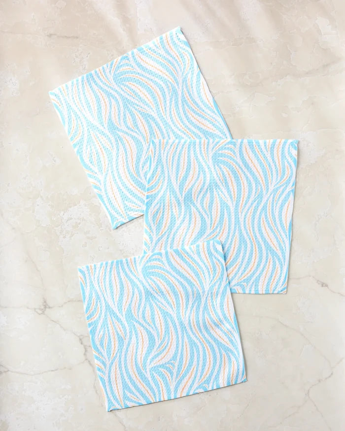 Three Rachel Parker Sky Willow Wave reusable cleaning cloths, featuring a blue and peach wavy pattern, are arranged in a loose triangle on a light marble surface.