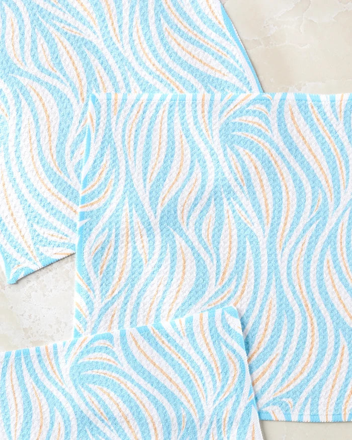 Three Sky Willow Wave dishcloths by Rachel Parker, featuring blue and white wavy patterns with touches of tan, are displayed on light marble. This super-absorbent set is made from recycled materials for eco-friendly cleaning.