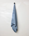 The GEOMETRY Sky waffle bath towel, in light blue, hangs neatly from a black hook on a white wall, bringing an elegant touch of simple luxury to the space.