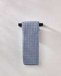 The GEOMETRY Sky hand towel, neatly folded in light blue waffle texture, hangs on a sleek black bar against a plain wall for an understated style touch.