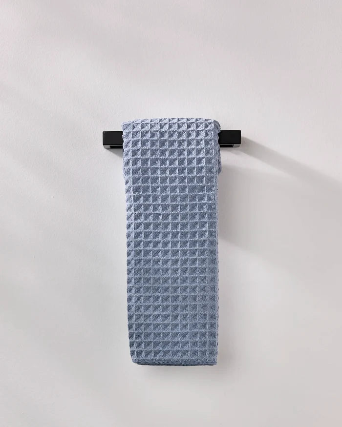 The GEOMETRY Sky hand towel, neatly folded in light blue waffle texture, hangs on a sleek black bar against a plain wall for an understated style touch.