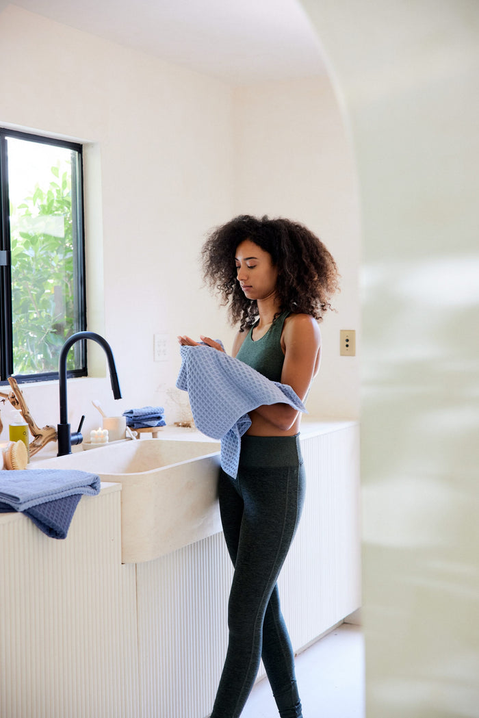 A woman with curly hair dries a dish by the sink in a modern kitchen using the ultra-absorbent GEOMETRY Sky waffle hand towel, sunlight streaming through the window and kitchen items neatly arranged on the counter.
