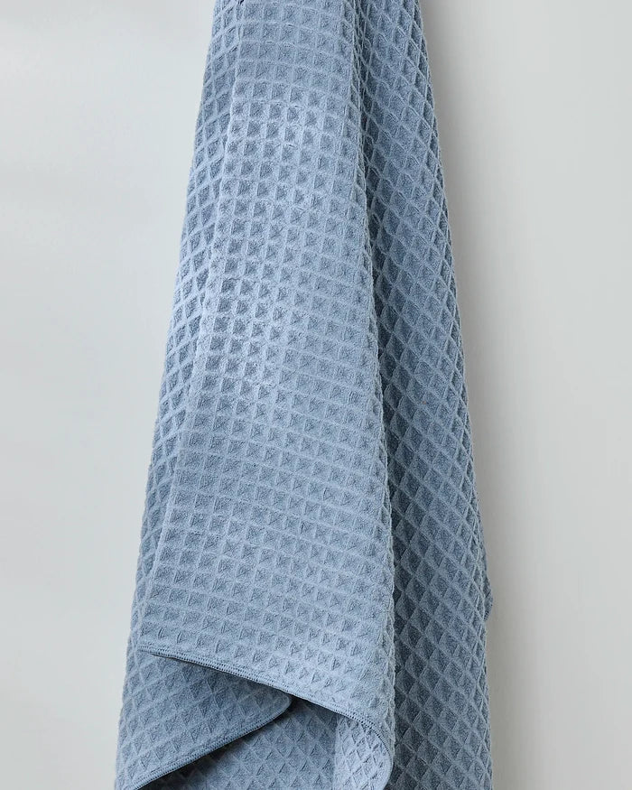 The GEOMETRY Sky bath towel, in light blue, hangs against a light gray wall, showing off its soft waffle texture and neatly draped fabric.