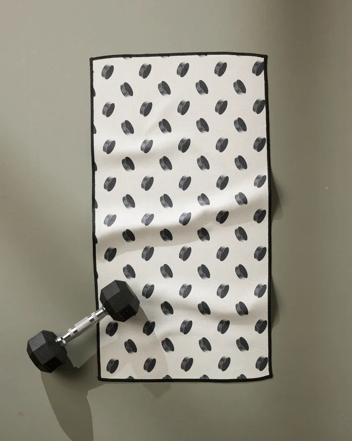 A Geometry Slapshot towel with a black dotted pattern lies next to a black dumbbell, both resting on a plain gray surface.