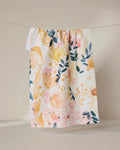 The GEOMETRY Sleepy Fields white kitchen towel, featuring a vibrant floral pattern in yellow, pink, and blue and made from recycled materials, hangs on a string against a neutral beige background.