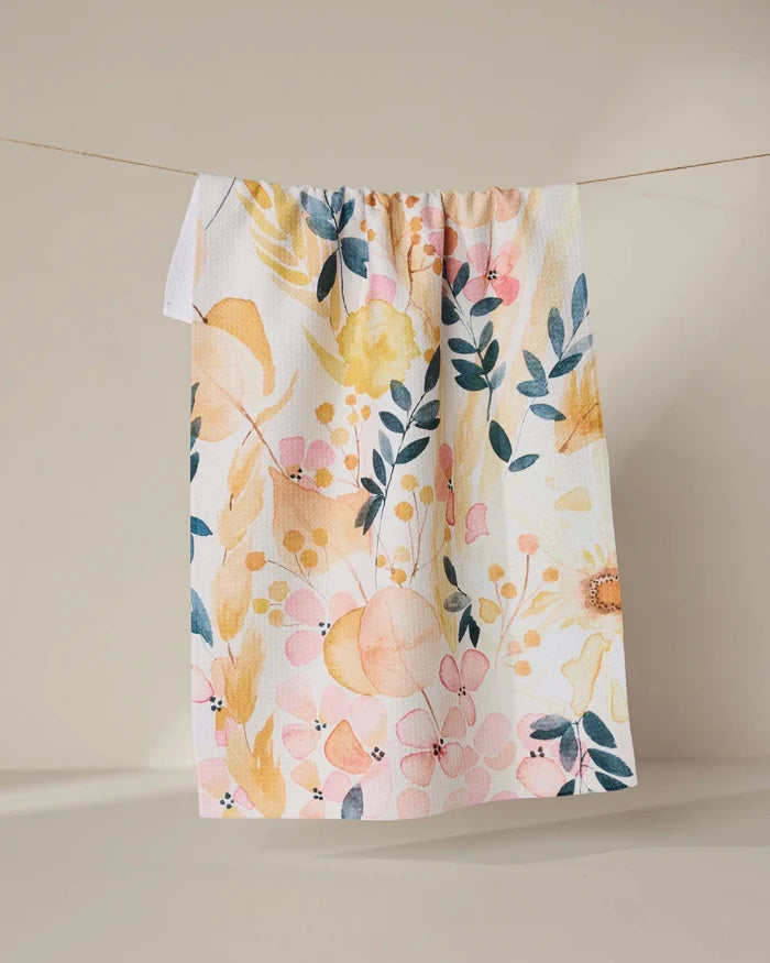 The GEOMETRY Sleepy Fields white kitchen towel, featuring a vibrant floral pattern in yellow, pink, and blue and made from recycled materials, hangs on a string against a neutral beige background.