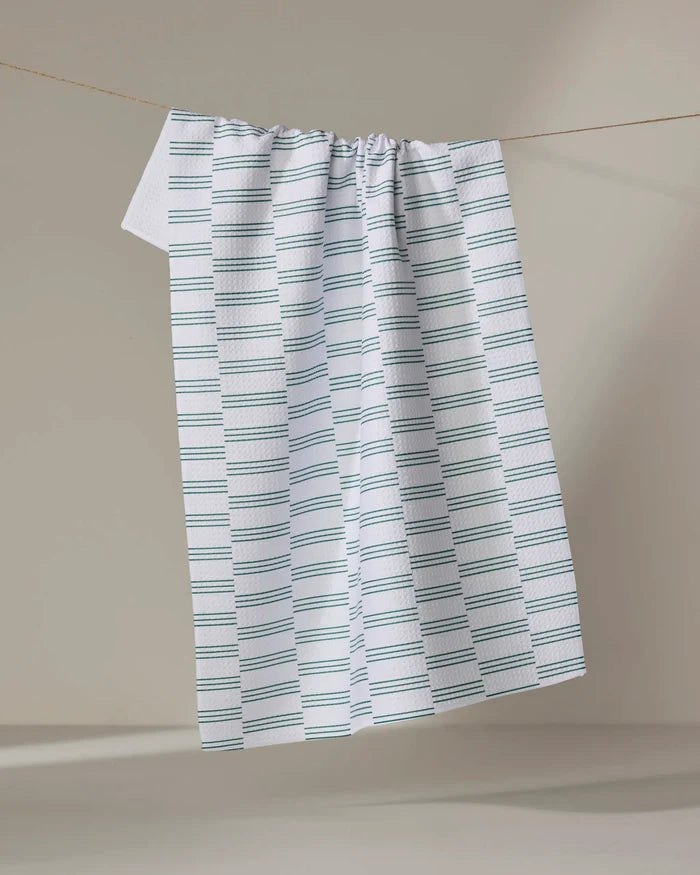 A Small Stripe - Evergreen dish towel by The Buy Guide, featuring blue and gray horizontal stripes, hangs on a clothesline against a beige background.