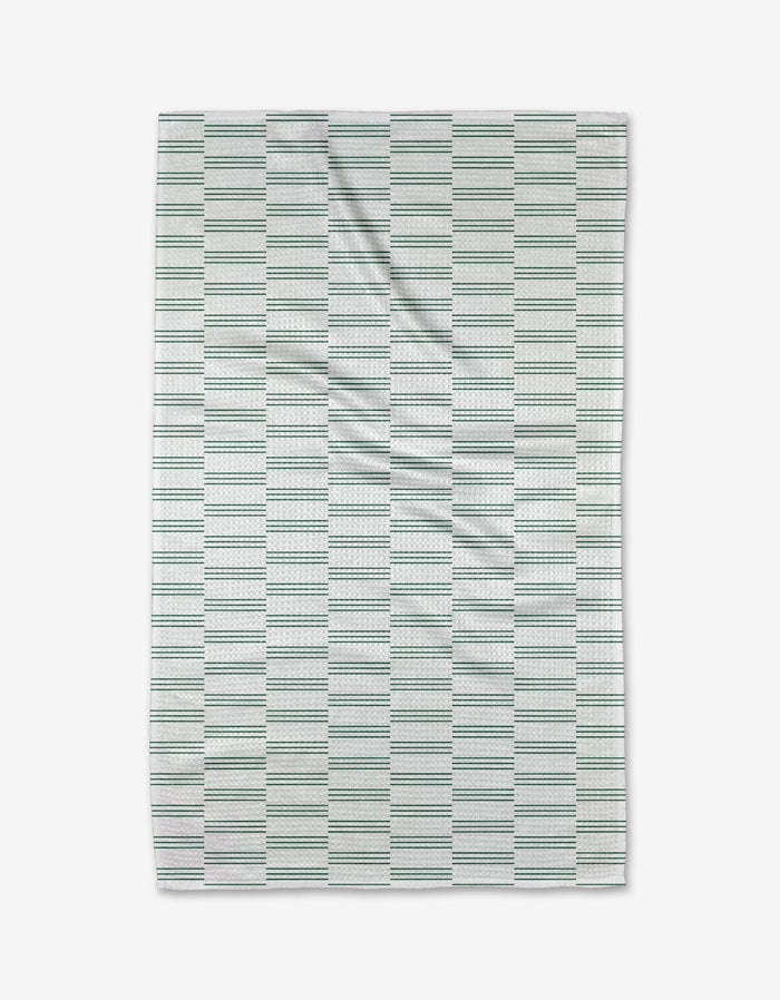 The Buy Guide’s Small Stripe - Evergreen tea towel features a white background with thin green and blue stripes in rectangular blocks. Crafted from Geoweave fabric, it’s pictured laid flat with subtle wrinkles.