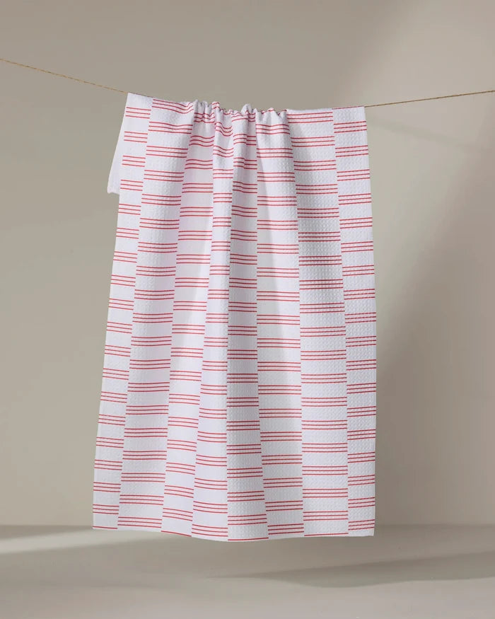 The Buy Guide's Small Stripe - Red tea towel, featuring evenly spaced horizontal red stripes on white, hangs from a string against a plain, neutral background.