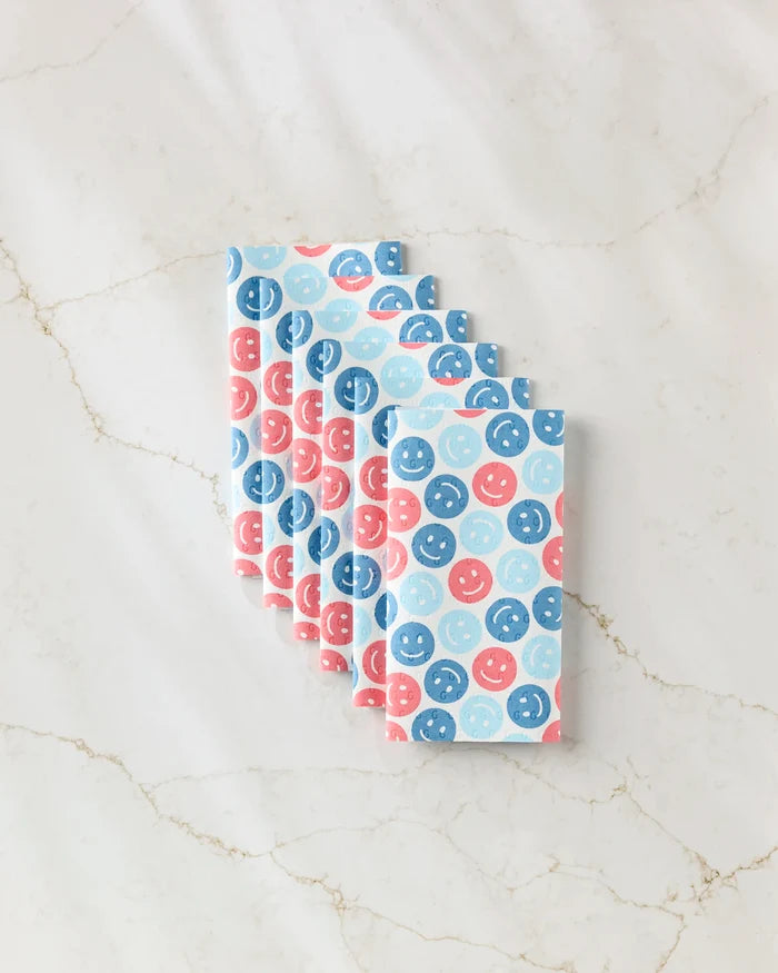 Six Hufton Studio "Smiley Face July Blue" folded reusable paper towels, featuring blue and pink smiley faces, are stacked neatly on a white marble surface with light brown veining.