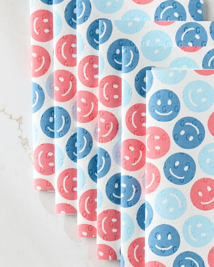 Five eco-friendly "Smiley Face July Blue" kitchen towels from Hufton Studio, featuring pink, red, and blue smiley faces, are neatly fanned out on a white marble surface.
