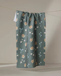 The Snail Gathering kitchen towel by Amberleigh Adoff, featuring beige snails, light blue flowers, and white geometric shapes on dark teal, hangs from a taut string against a neutral wall.