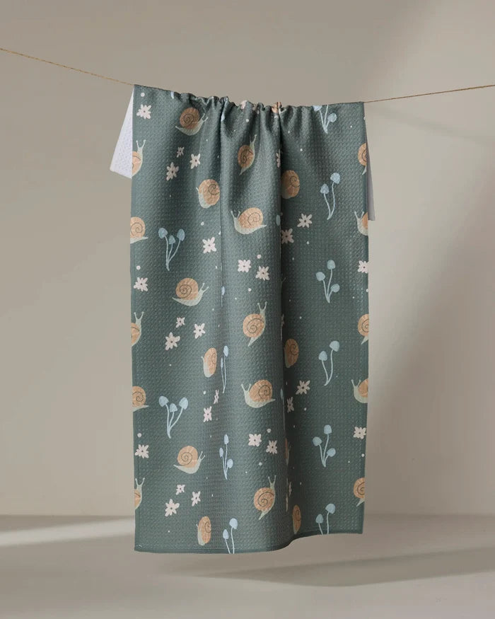 The Snail Gathering kitchen towel by Amberleigh Adoff, featuring beige snails, light blue flowers, and white geometric shapes on dark teal, hangs from a taut string against a neutral wall.