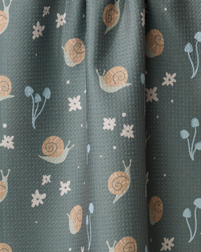 A close-up of the Snail Gathering dish towel by Amberleigh Adoff shows its absorbent green fabric with a playful print of snails, white flowers, and blue mushrooms.