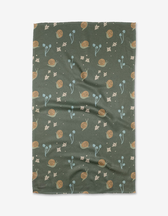 The Snail Gathering tea towel by Amberleigh Adoff is green and textured, made from recycled materials, and features brown snails, blue plants, and cream flowers. Shown flat with subtle wrinkles on a white background.