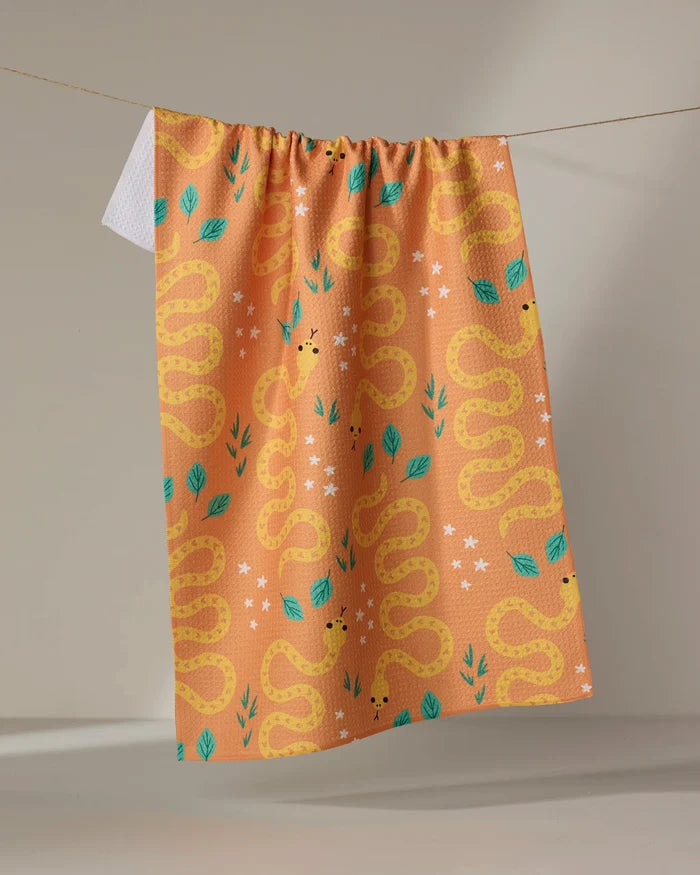 The Genna Blackburn "Snakes" tea towel in peach features a repeating yellow snake pattern with green leaves and white star-shaped flowers and is ultra-absorbent, perfect for hanging on your line.