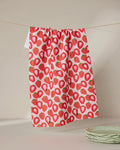 The Geometry So Many Strawberries tea towel, made from quick-drying recycled materials, hangs on a clothesline against a neutral background, with light green scalloped plates stacked in the lower right corner.