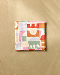 The Soft Cutouts Garden beach blanket by Claire Iglesias is a square, quick-drying piece made from recycled materials with an abstract geometric pastel design in orange, green, pink, and purple, shown folded on a neutral beige surface.