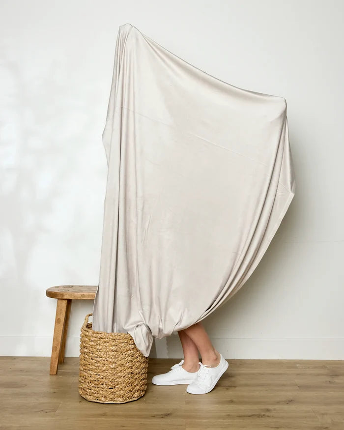 A person stands on a woven basket with their legs visible, wrapped in a GEOMETRY Soft Glow throw blanket, next to a wooden stool in a room with light-colored walls and a wooden floor.