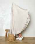 A person stands on a woven basket with their legs visible, wrapped in a GEOMETRY Soft Glow throw blanket, next to a wooden stool in a room with light-colored walls and a wooden floor.
