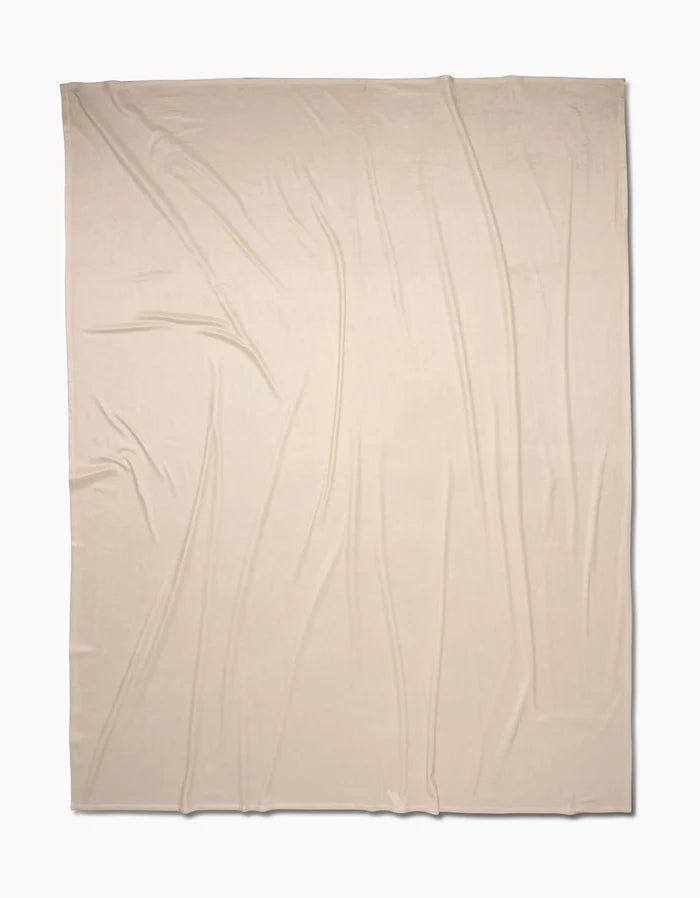 A light beige, rectangular Soft Glow blanket by GEOMETRY is displayed flat with gentle, natural folds. Made from eco-friendly recycled materials, the fabric rests on a plain white background.