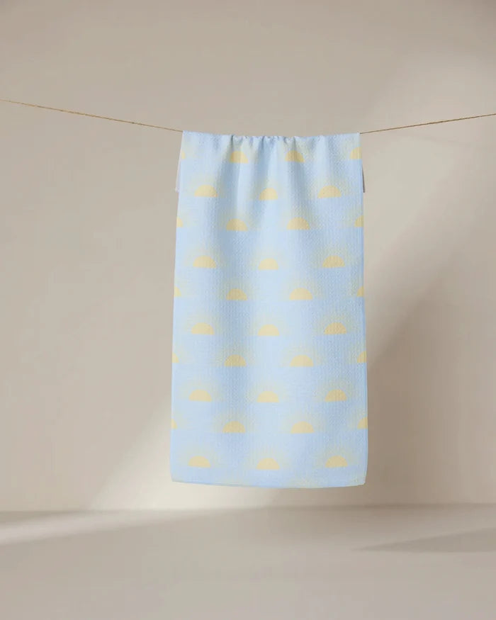 The GEOMETRY Soft Summer Sun towel, ultra-absorbent and light blue with a pattern of yellow semicircles, hangs on a clothesline against a softly lit neutral background.