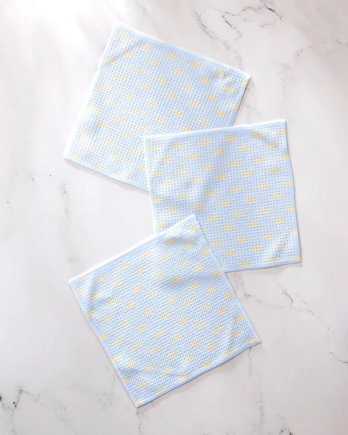 Three GEOMETRY "Soft Summer Sun" light blue textured recycled dishcloths with small yellow dots are arranged on a white marble surface.