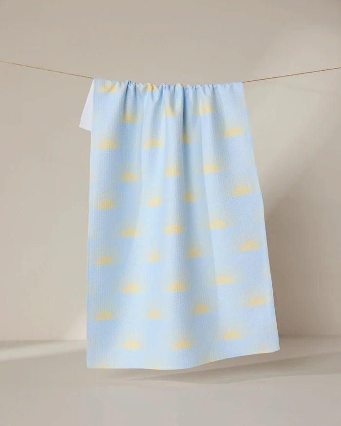 The Soft Summer Sun tea towel by GEOMETRY, made from recycled materials and decorated with yellow semicircles on pastel blue, hangs from a clothesline against a neutral background.