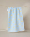 The Soft Summer Sun tea towel by GEOMETRY, made from recycled materials and decorated with yellow semicircles on pastel blue, hangs from a clothesline against a neutral background.