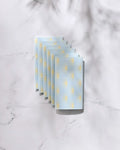 Six folded GEOMETRY Soft Summer Sun blue reusable paper towels with yellow polka dots are arranged in a neat, overlapping row on a white marble surface with soft shadows.