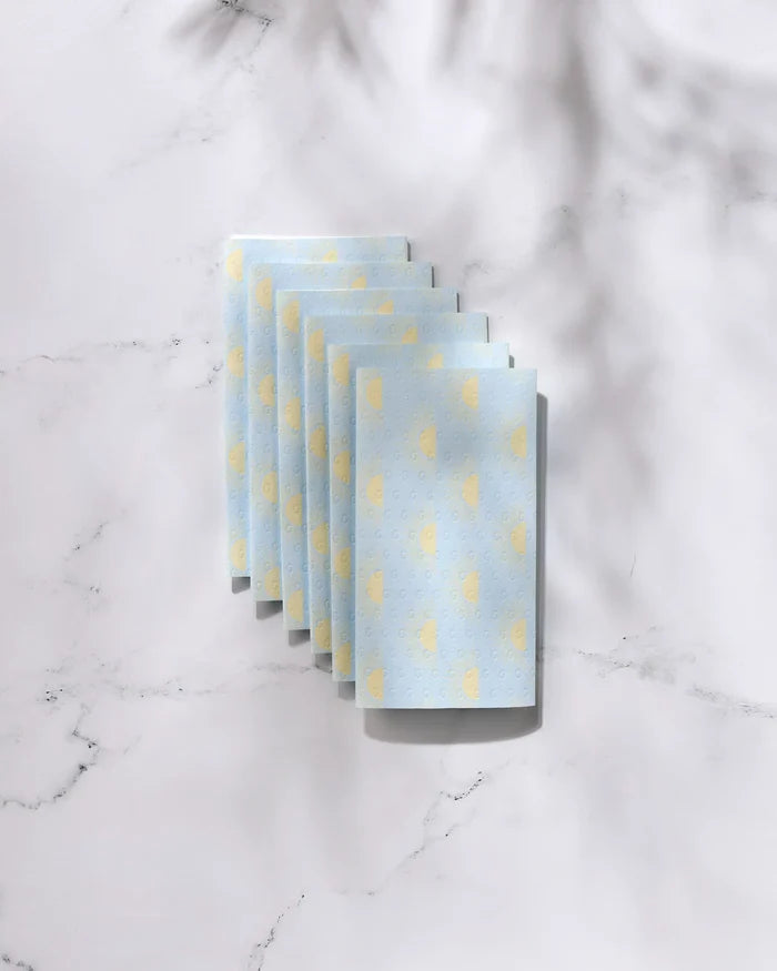 Six folded GEOMETRY Soft Summer Sun blue reusable paper towels with yellow polka dots are arranged in a neat, overlapping row on a white marble surface with soft shadows.
