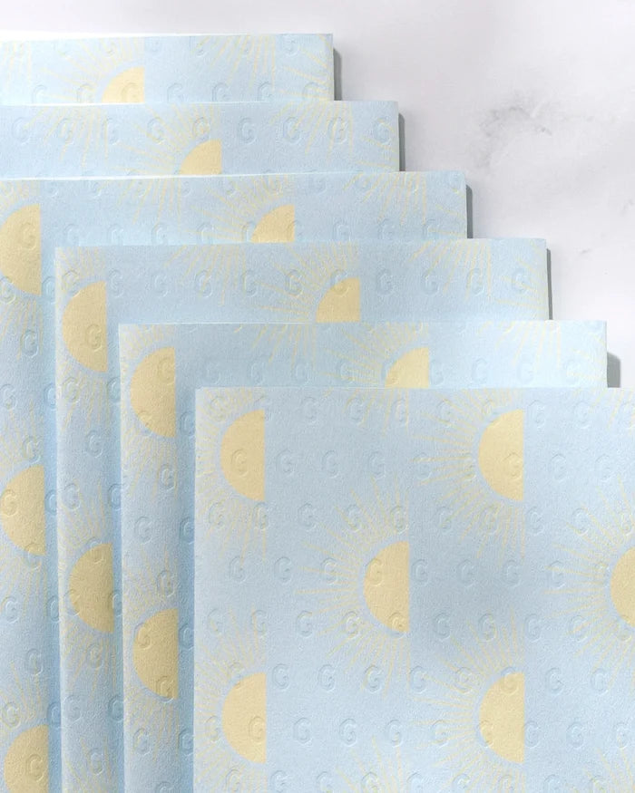 Several sheets of GEOMETRY's Soft Summer Sun eco-friendly, light blue paper towels—decorated with yellow suns and a subtle “G” emboss—are arranged in a staggered, overlapping layout on a light background.