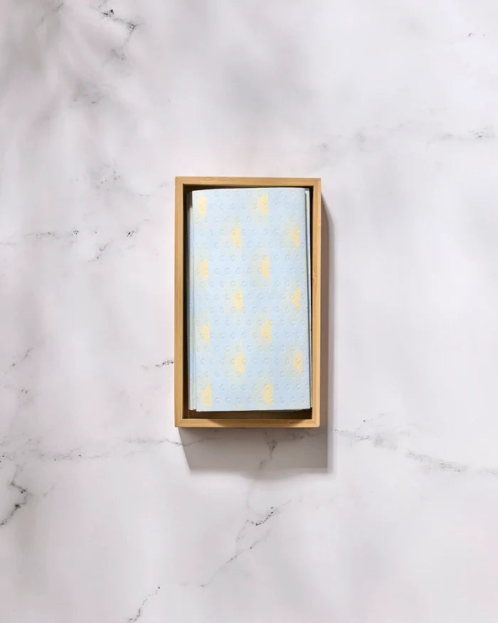 A rectangular wooden tray holds GEOMETRY's Soft Summer Sun kitchen towel, featuring a subtle yellow pattern on light blue, displayed on white marble.