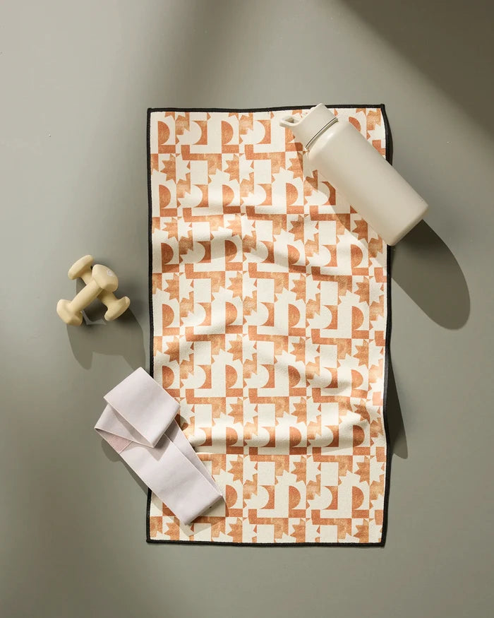 The Geometry Solar Power fitness towel is laid flat on a gray surface, with a white water bottle, cream dumbbells, and a folded white strap placed on and beside it. The towel features an eye-catching pattern and is made from recycled materials.