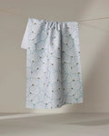 The Solie Teal tea towel by Amy MacCready, crafted from recycled materials, features a quick-drying white fabric with a light blue scallop shell pattern and small yellow bee accents, hanging against a neutral background.