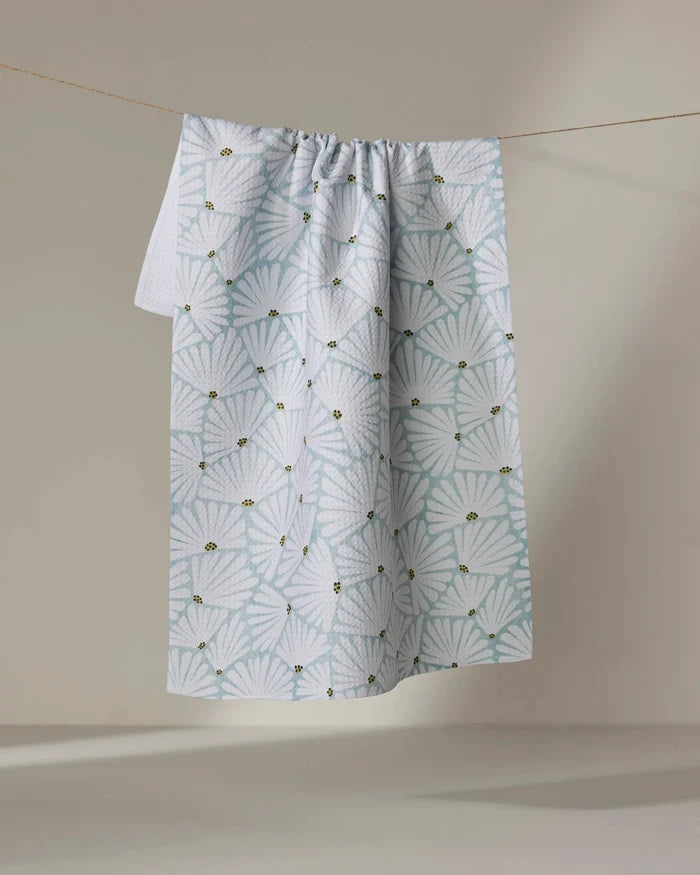 The Solie Teal tea towel by Amy MacCready, crafted from recycled materials, features a quick-drying white fabric with a light blue scallop shell pattern and small yellow bee accents, hanging against a neutral background.