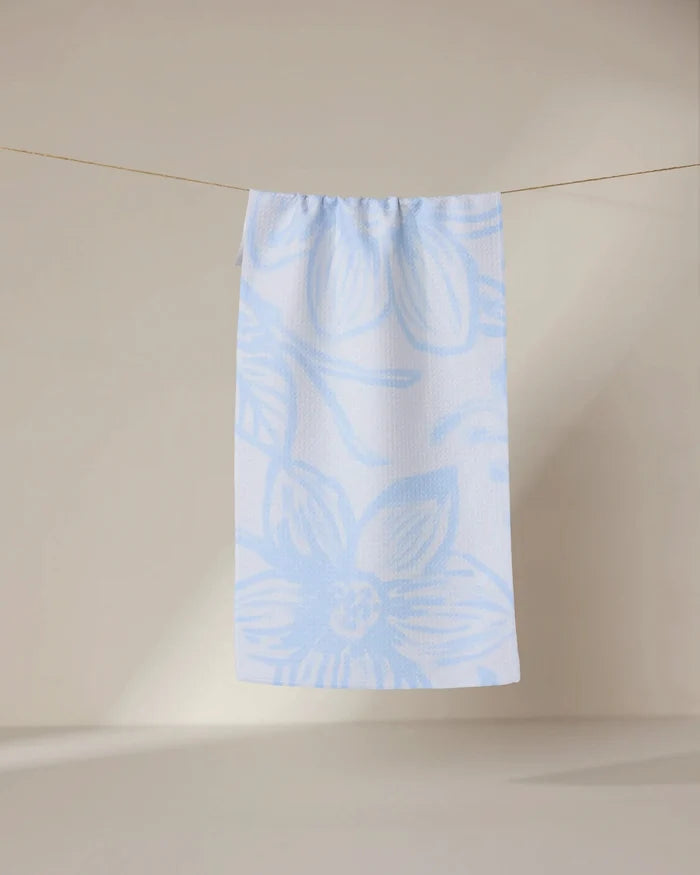 The GEOMETRY Something Blue bar towel, featuring light blue floral patterns, hangs on a thin clothesline against a beige background with soft light casting gentle shadows on the wall and floor.
