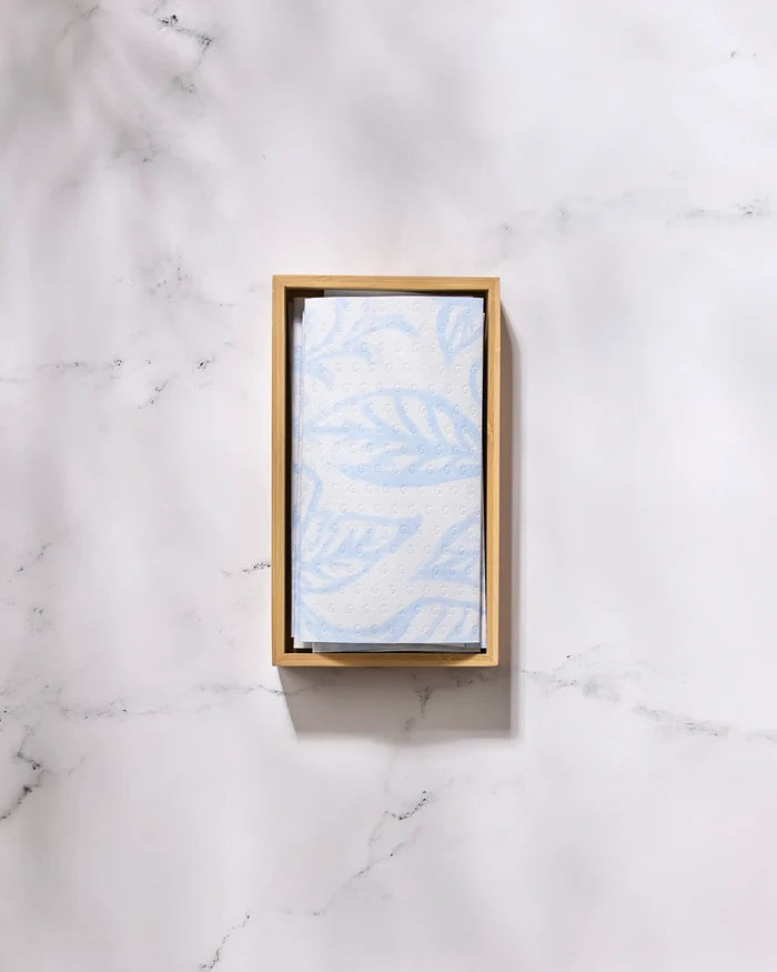 The GEOMETRY Something Blue eco-friendly, light blue paper towel with a leaf pattern is folded in a rectangular wooden tray on a white marble surface.