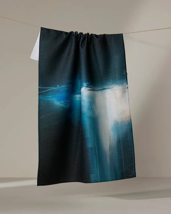 The “Somewhere” tea towel by Jennifer Janesko, with an abstract dark blue, teal, and white brushstroke design, hangs on a line against a neutral background.