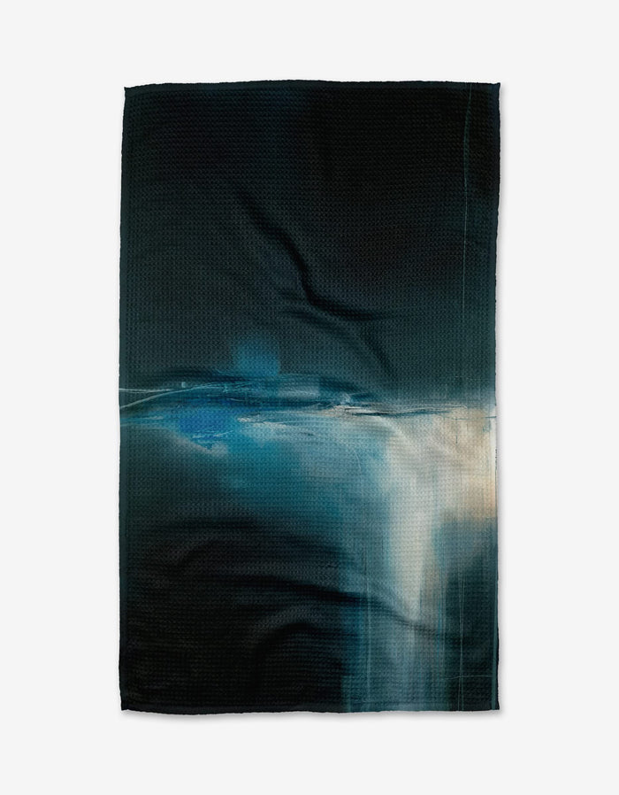 The Jennifer Janesko Somewhere tea towel is dark, textured, and made from recycled materials, featuring a blue and white abstract pattern inspired by light on water, shown on a plain white background.