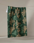 The Geometry Sonnet Forest tea towel, featuring a dark green floral pattern with tan and light green accents, hangs draped over a string against a neutral background.