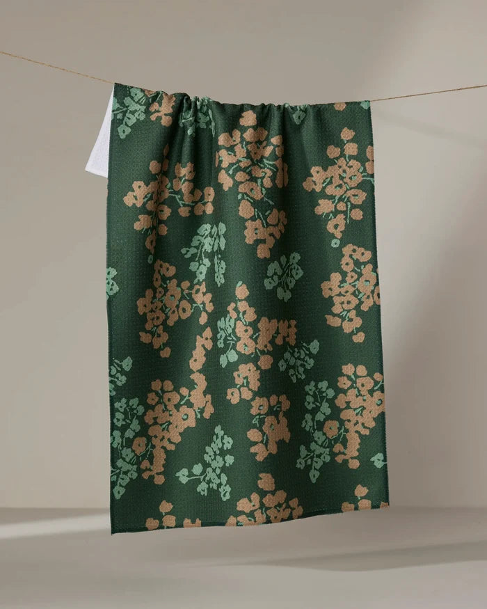 The Geometry Sonnet Forest tea towel, featuring a dark green floral pattern with tan and light green accents, hangs draped over a string against a neutral background.