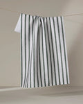 A Becki Owens "Spearmint" tea towel made of white Geoweave fabric with vertical dark green stripes hangs draped on a string, forming gentle folds against a light beige background.