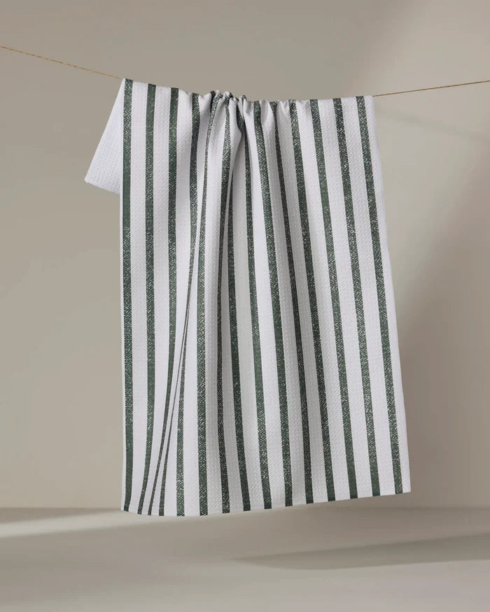 A Becki Owens "Spearmint" tea towel made of white Geoweave fabric with vertical dark green stripes hangs draped on a string, forming gentle folds against a light beige background.