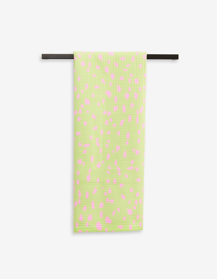 The GEOMETRY Speckle Green tea towel, featuring light green Geoweave fabric with pink spots, hangs neatly on a black towel bar against a white background.